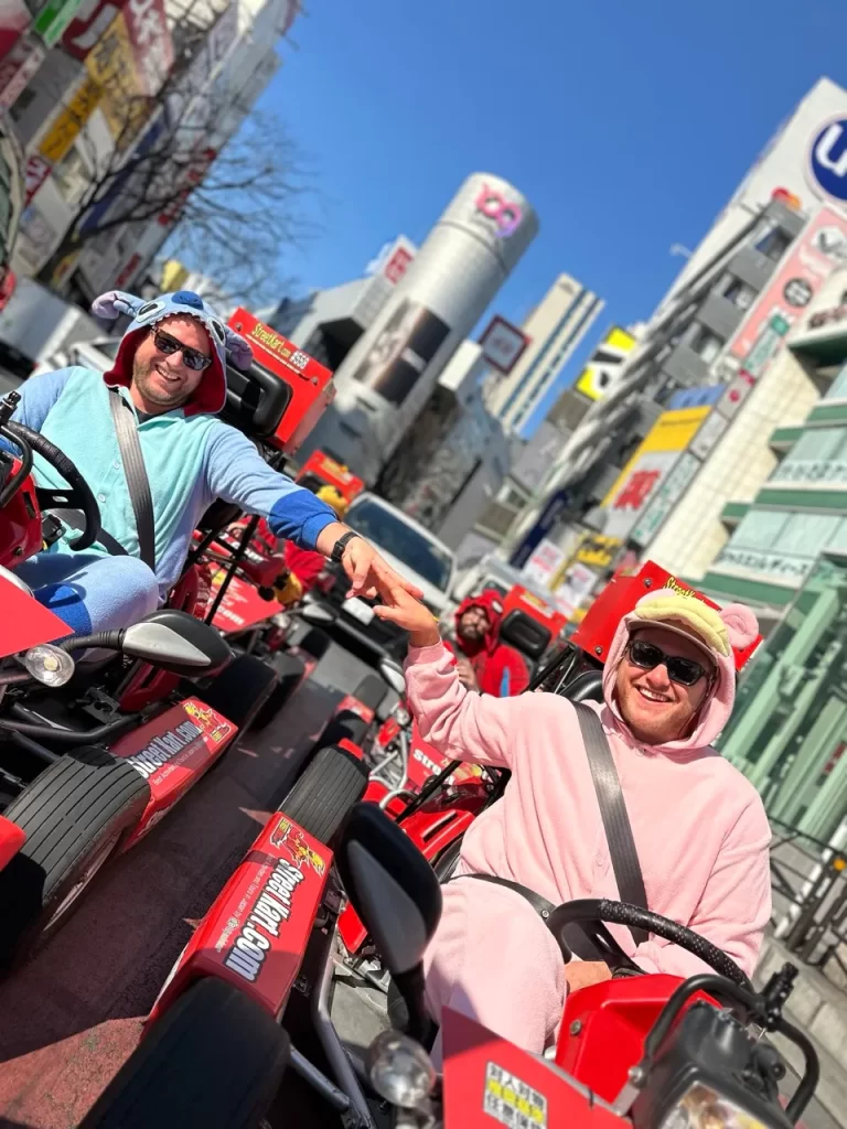 Two men in colorful hoodies riding red go-karts on a busy city street, giving a high-five and smiling at the camera, with tall buildings in the background.
