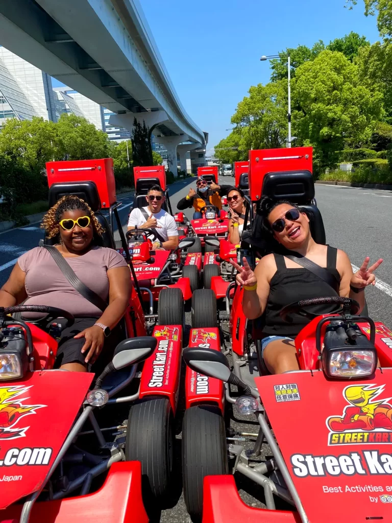 Group of people in bright red go-karts on a sunny street, smiling and posing for the camera under an elevated roadway.