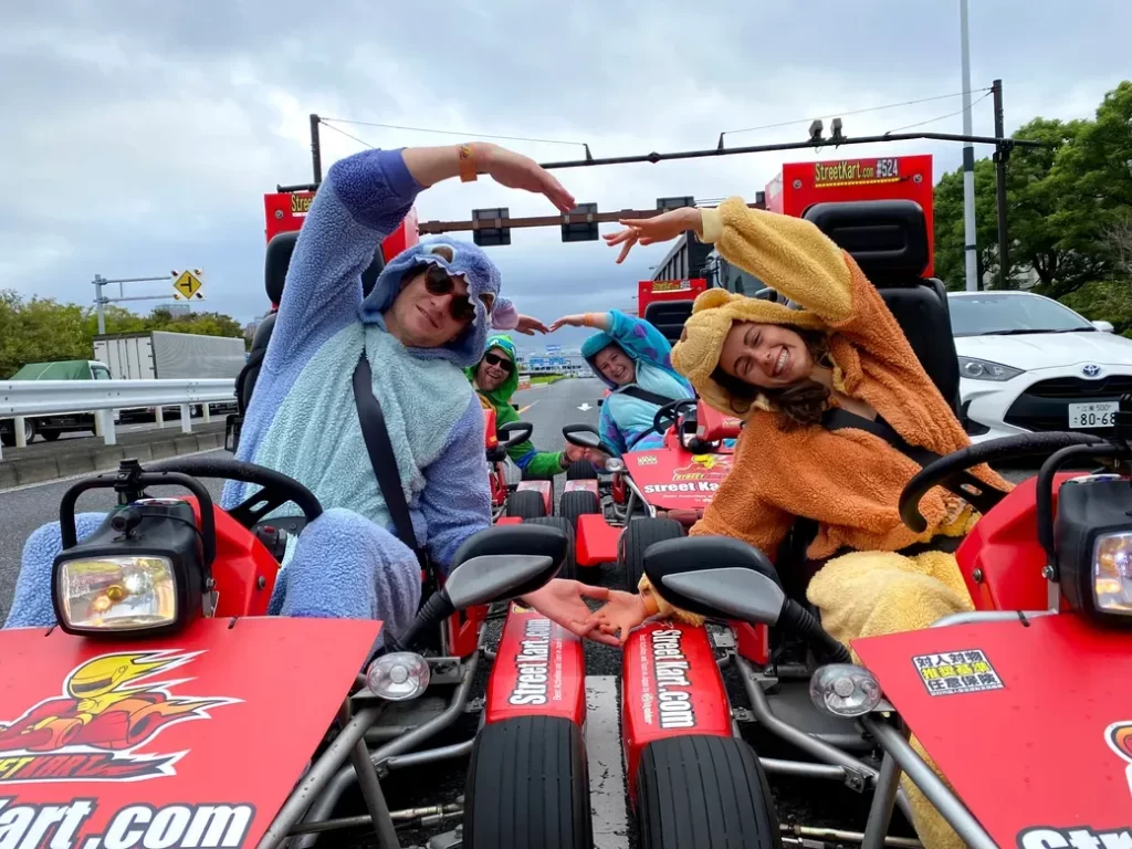 Group of people in colorful plush costumes on go-karts forming a heart shape with their arms.